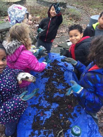 Tilth Alliance facilitates project-based learning with South Shore Students. Six children crowd around a blue tarp with dirt and critters on it. The children are in winter clothes.
